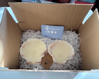 Puede incluir: Una caja de cartón marrón rellena de papel blanco triturado, que contiene dos velas de color blanco roto con forma de concha. Una tarjeta rectangular azul claro dice Birthday Beach Box con tres iconos de flores. Una pequeña etiqueta marrón dice made with y un símbolo de corazón.