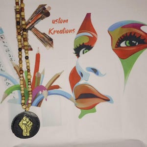 May include: A wooden beaded necklace with a black pendant featuring a gold glitter fist. The background includes a colorful abstract face design and the text "ustom Kreations". Pencils and art supplies are also visible.
