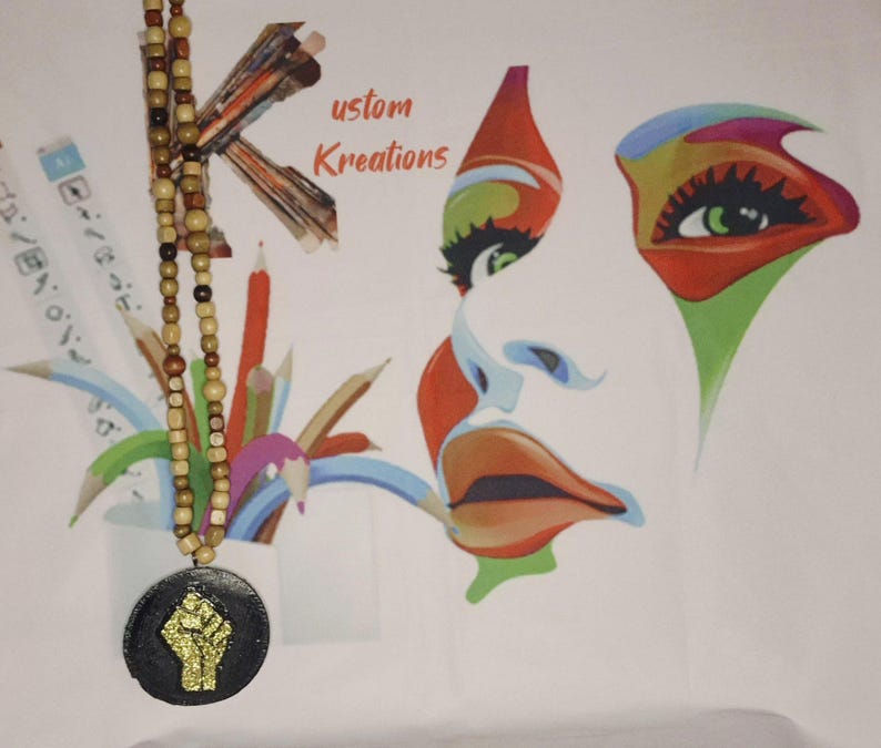 May include: A wooden beaded necklace with a black pendant featuring a gold glitter fist. The background includes a colorful abstract face design and the text "ustom Kreations". Pencils and art supplies are also visible.