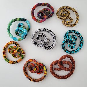 May include: Eight fabric-covered pretzel-shaped ornaments in various patterns and colors, including leopard print, black and white geometric, solid brown, and multi-colored floral and abstract designs, arranged in a circular formation.