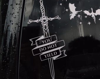 May include: White vinyl sticker featuring a sword design with the words "YOU DO NOT YIELD" on a banner. The sticker also includes silhouettes of dragons and stars, set against a black background.