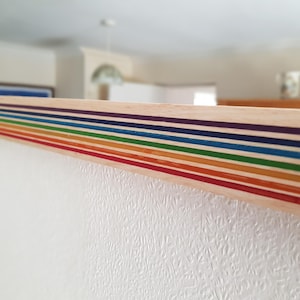 Pode incluir: Uma moldura decorativa de madeira com listras de arco-íris. A moldura apresenta linhas paralelas em vermelho, laranja, amarelo, verde, azul, índigo e violeta. É montada em uma parede branca texturizada. A moldura tem cerca de 100 cm de comprimento.