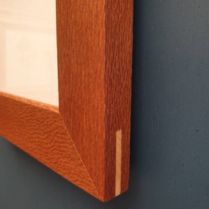 May include: Close-up of a wooden picture frame with a mitered corner. The frame is made of a reddish-brown wood with a natural finish. The corner is joined with a small piece of wood that is a lighter color.
