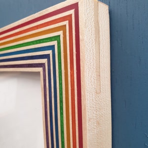 May include: Close-up of the corner of a rainbow-colored wooden picture frame. The frame is made of thin strips of wood, each painted a different color of the rainbow. The frame is set against a blue background.