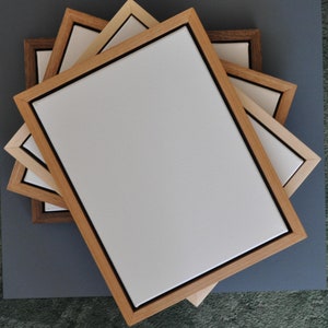 May include: Three wooden picture frames of different sizes and colours, stacked on top of each other. The frames are made of natural wood with a black border around the opening. The frames are empty and ready for a picture.