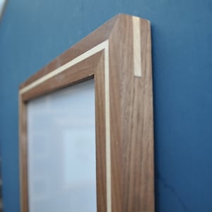 May include: A close-up of a wooden picture frame with a light wood inlay. The frame is made of walnut wood and has a simple, modern design.