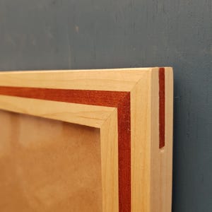 Puede incluir: Primer plano de un marco de fotos de madera con un borde de madera clara y una fina franja roja oscura. El marco está sobre un fondo verde azulado oscuro. Se ve la esquina del marco.