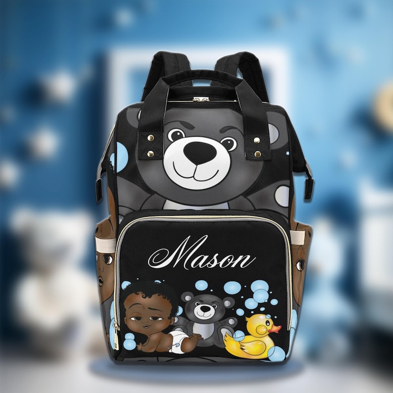 May include: Black and gray diaper bag with a bear face design on the top. The front pocket features the name "Mason" in white script, along with illustrations of a baby, a teddy bear, and a rubber ducky. The bag has multiple pockets and adjustable straps.