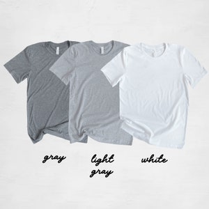 May include: Three t-shirts in different colors: gray, light gray, and white. The t-shirts are laid out flat on a white surface.