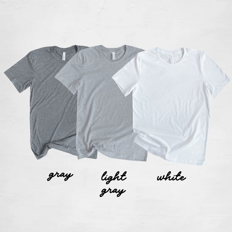 May include: Three t-shirts in different colors: gray, light gray, and white. The t-shirts are laid out flat on a white surface.