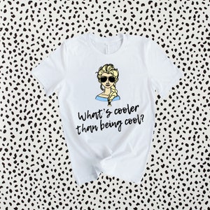 May include: White t-shirt with a cartoon image of a woman with blonde hair wearing sunglasses and a blue dress. The text "What's cooler than being cool?" is printed below the image.