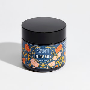 Puede incluir: Un tarro negro con una etiqueta azul y dorada con un diseño floral. La etiqueta dice "Remnant Beauty Tallow Balm" y "Net Wt. 60ml".