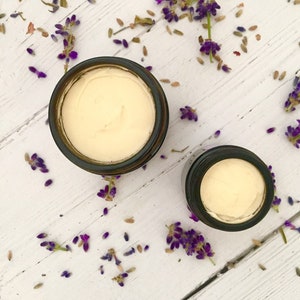 May include: Two glass jars with lids containing a white cream, surrounded by lavender flowers on a white wooden surface.