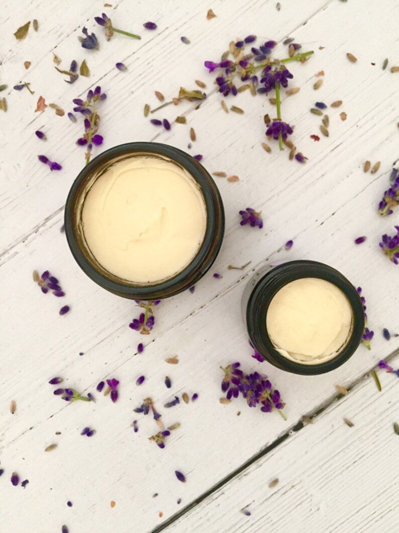 May include: Two glass jars with lids containing a white cream, surrounded by lavender flowers on a white wooden surface.