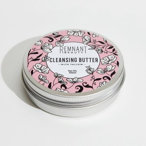 May include: A silver tin with a pink label featuring a floral design. The label reads "Remnant Beauty Cleansing Butter - With Tallow - Net Wt. 100ml".
