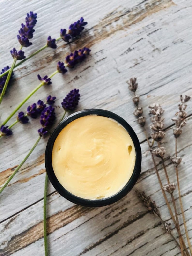 May include: A small black jar filled with a pale yellow cream, surrounded by sprigs of lavender flowers on a white wooden surface.