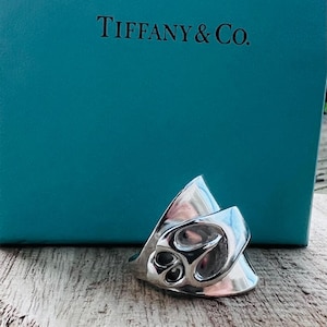 May include: A silver ring with a unique, abstract design. The ring is sitting on a wooden surface in front of a Tiffany & Co. blue box.