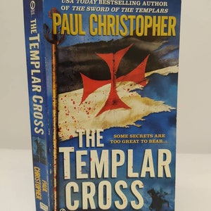 Puede incluir: Una portada de libro de bolsillo con un fondo azul y una cruz roja sobre una bandera blanca. El título del libro es "The Templar Cross" de Paul Christopher. El texto "Some secrets are too great to bear..." también está en la portada.