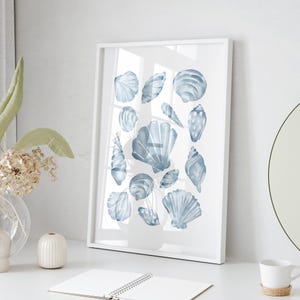 Puede incluir: Una impresi&oacute;n enmarcada que presenta una colecci&oacute;n de conchas marinas en acuarela. Las conchas, de diversas formas y tama&ntilde;os, est&aacute;n pintadas en tonos azules y grises sobre un fondo blanco. La obra de arte se presenta en un marco blanco, ideal para decoraci&oacute;n costera.