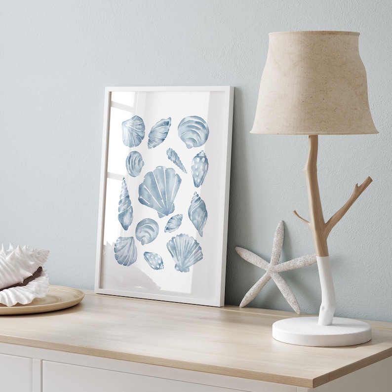 Puede incluir: Un cuadro enmarcado con ilustraciones de acuarela de conchas marinas azules. Una pantalla de l&aacute;mpara beige se encuentra sobre una l&aacute;mpara con forma de rama de &aacute;rbol. Una estrella de mar blanca decora la superficie de madera.