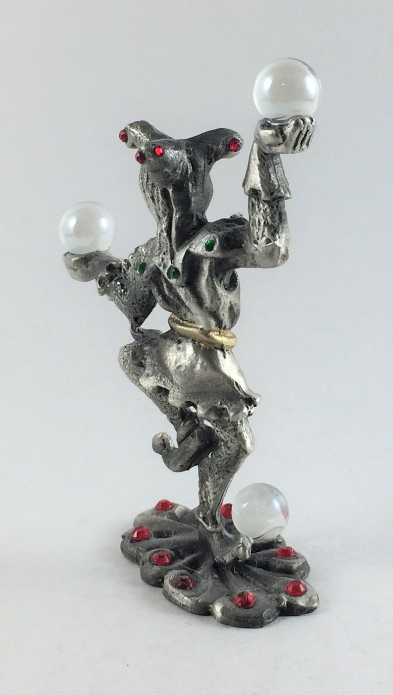May include: A silver-toned figurine of a jester holding three clear orbs. The jester has red and green accents, a gold ring around the waist, and a decorative base with red gems. A unique decorative item.