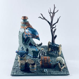 May include: Miniature fantasy diorama featuring a forge with a blue and gray structure, an anvil, barrels, a tree, and a figure. The scene includes a chest, a grindstone, and a small figure with wings. The base is a gray cobblestone.