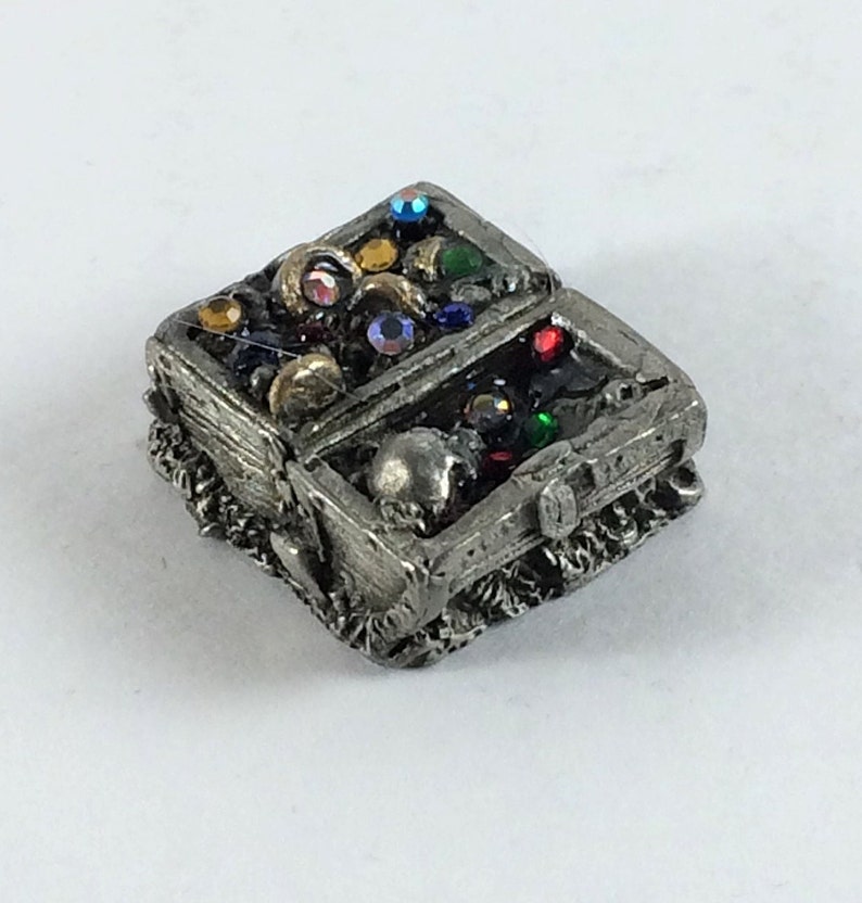 May include: A small pewter treasure chest with a skull on the lid, open to reveal a variety of colorful gems and jewels.