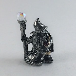 May include: A pewter figurine of a wizard holding a staff with a crystal ball on top. The wizard is wearing a pointed hat and a long robe. The figurine is painted in silver and gold.