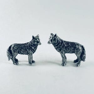 May include: Two small, detailed silver-colored wolf figurines. The wolves are standing, facing each other, with textured fur and realistic features. The figurines are on a white background.