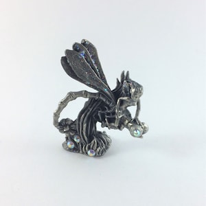 May include: A pewter figurine of a fairy with iridescent wings perched on a tree stump. The fairy is holding a small, round object in its hand. The figurine is decorated with iridescent rhinestones.