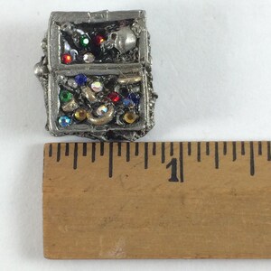May include: A small, silver-colored treasure chest with a skull and crossbones design. The chest is filled with colorful jewels and gems.