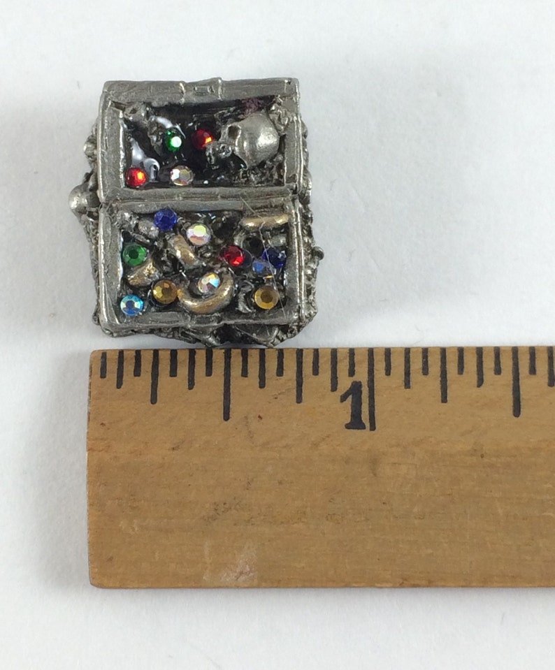 May include: A small, silver-colored treasure chest with a skull and crossbones design. The chest is filled with colorful jewels and gems.