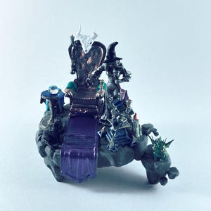 May include: A miniature diorama featuring a dark throne with a white skull topper, a figure in a black pointed hat, a small blue table with a crystal ball, a purple rug, and a small green spiky creature on a rocky gray base.