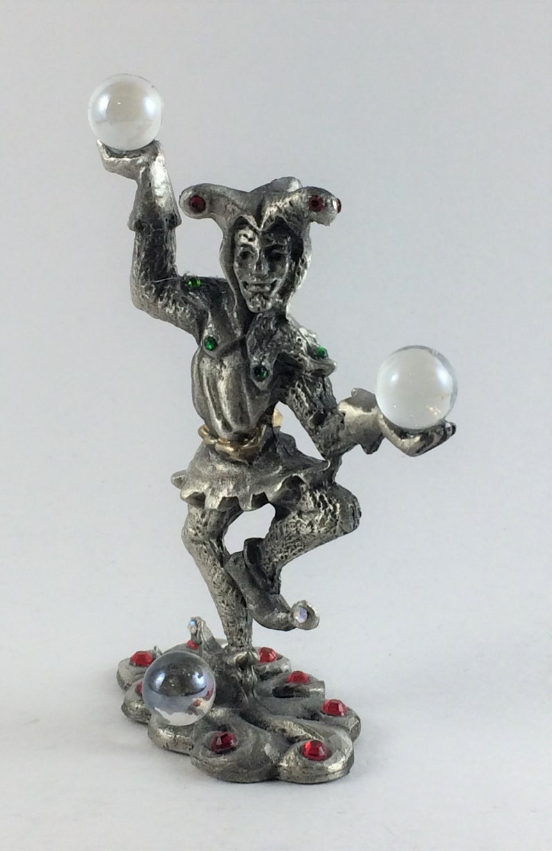 May include: A silver-colored figurine of a jester balancing two clear orbs. The jester wears a hat with red accents and has green and red gems on the body. The base is decorated with red gems and a clear orb.