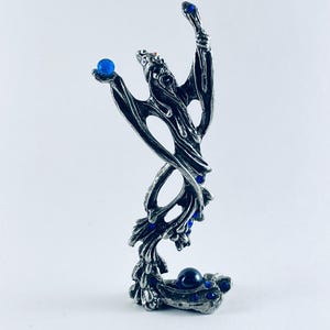 May include: A silver-toned figurine of a stylized figure with outstretched arms, holding a blue orb. The figure is adorned with several blue gemstones. The base of the figurine also features a blue orb and additional blue gemstones.