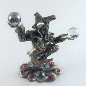 May include: A silver-colored figurine of a jester holding two crystal balls. The jester is adorned with red and green jewels and stands on a cloud-shaped base. The jester's hat has red jewels. The figurine is a decorative item.