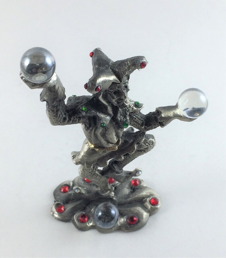 May include: A silver-colored figurine of a jester holding two crystal balls. The jester is adorned with red and green jewels and stands on a cloud-shaped base. The jester's hat has red jewels. The figurine is a decorative item.