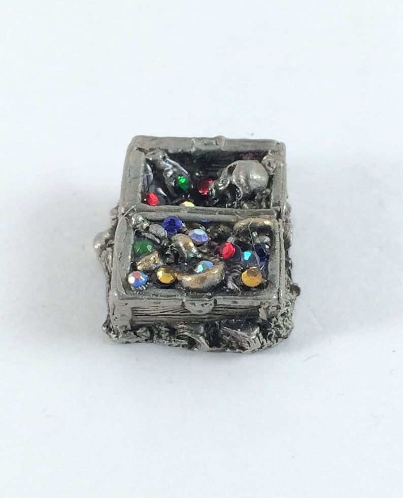 May include: A pewter treasure chest filled with colorful gems and a skull. The chest has a detailed design and is a miniature collectible.