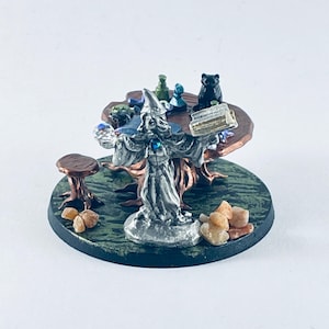 May include: A miniature silver-colored wizard figurine wearing a pointed hat and robes, holding an open book and a crystal. The figure stands on a round base with small rocks, a wooden table with various small items, and a stool.