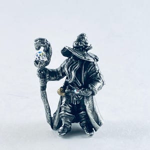 May include: A detailed miniature figurine of a wizard, crafted in silver-toned metal. The wizard wears a long coat and hat, holding a staff with a crystal ball. The figurine is standing on a white surface.