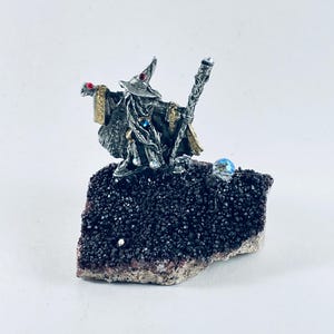 May include: A silver-toned wizard figurine wearing a pointed hat and robes, holding a staff and a small red-accented object. The figure is mounted on a dark, textured, crystalline mineral base with a single iridescent blue bead.
