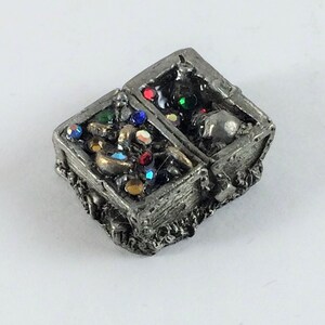 May include: A pewter treasure chest with a hinged lid open, revealing a collection of gold coins, jewels, and a skull inside. The chest is decorated with intricate details and has a rustic, aged appearance.