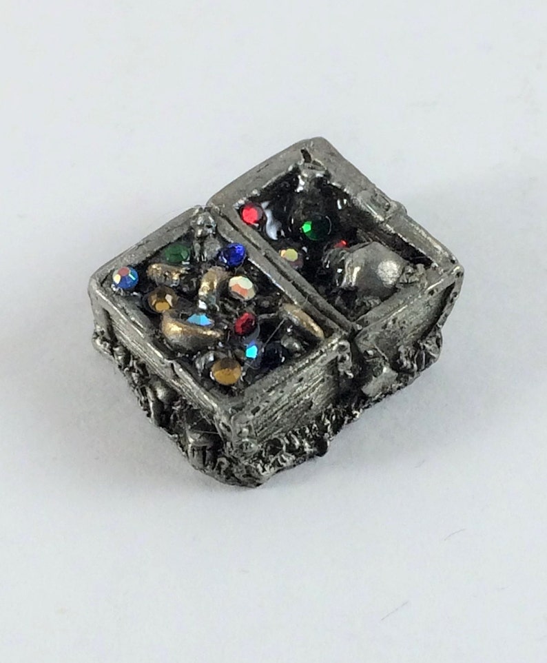 May include: A pewter treasure chest with a hinged lid open, revealing a collection of gold coins, jewels, and a skull inside. The chest is decorated with intricate details and has a rustic, aged appearance.