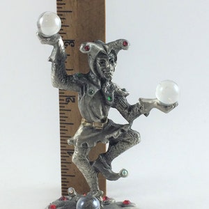 May include: A silver-colored figurine of a jester juggling two clear glass balls. The jester wears a hat with red accents and is adorned with red and green jewels. The base features a gray orb and red jewels. The figurine is approximately 4 inches tall.