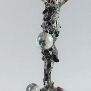 May include: A silver-colored decorative figurine with a textured surface. The figurine features a stylized design with three clear orbs and red and green accents. The base has a leaf-like shape with red embellishments.