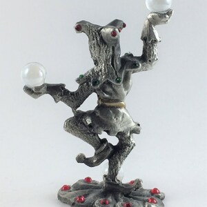 May include: A silver-colored figurine of a jester balancing two clear orbs. The jester is adorned with red and green jewels. The base of the figurine is decorated with red jewels.