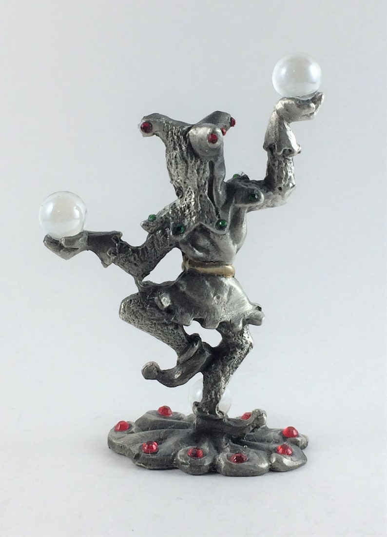 May include: A silver-colored figurine of a jester balancing two clear orbs. The jester is adorned with red and green jewels. The base of the figurine is decorated with red jewels.