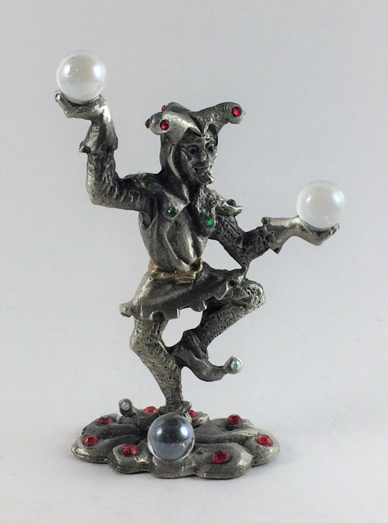 May include: A silver-colored figurine of a jester balancing two white spheres. The jester is adorned with red and green gemstones and stands on a cloud-shaped base with more red gems and a dark sphere. The jester's hat has three points.