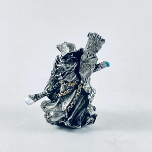 May include: A detailed silver-colored figurine of a robed wizard holding a staff with a crystal ball and a scroll. The wizard wears a hat and is adorned with a gold chain. A small blue gem is on the scroll.