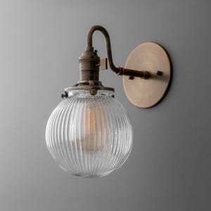 May include: A vintage-style wall sconce with a clear, ribbed glass globe. The fixture has a brass finish, including the curved arm, bulb socket, and circular wall mount. An Edison-style bulb is visible inside the globe.